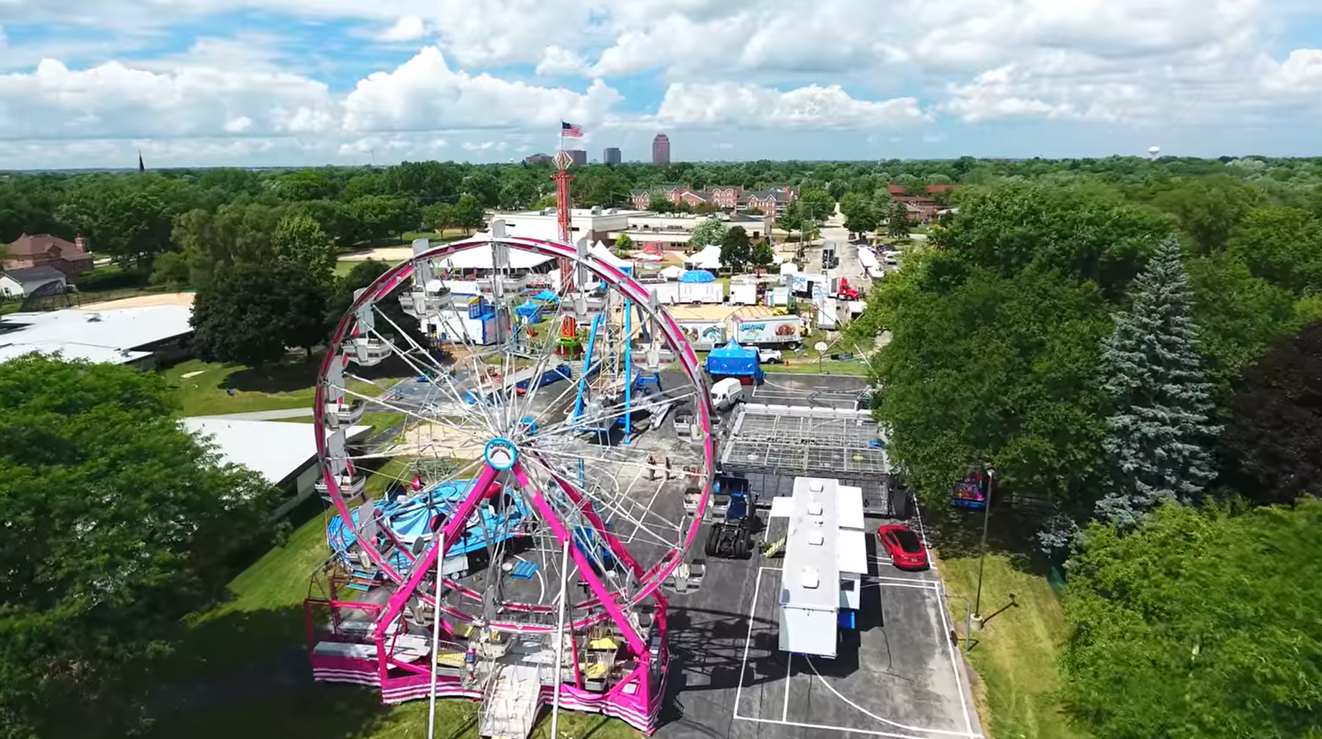 aerial shot of Ferris wheel, with adjacent carnival setups, surrounded by trees under a clear blue sky in the Itasca Fest area.