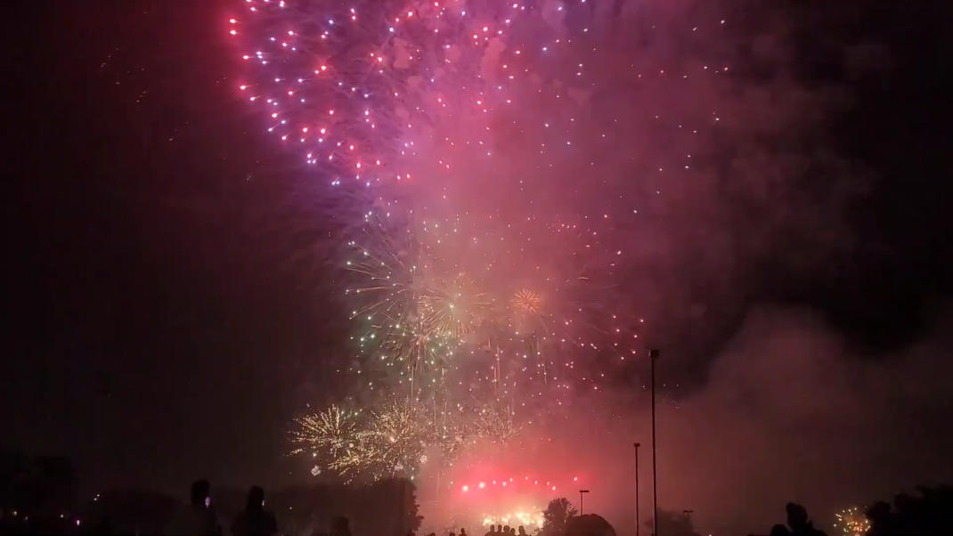 An explosion of pink and gold fireworks dazzles against a smoky backdrop, with silhouettes of spectators in the foreground witnessing the spectacle.