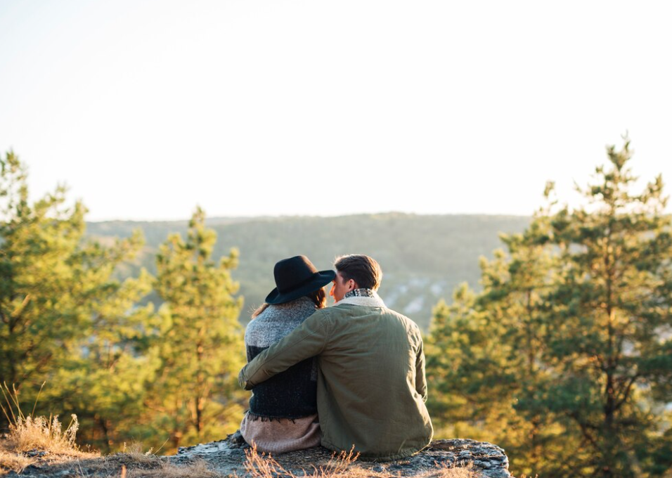A couple sits closely on a rocky outcrop, embracing each other and gazing at the forested valley below during a serene sunset.