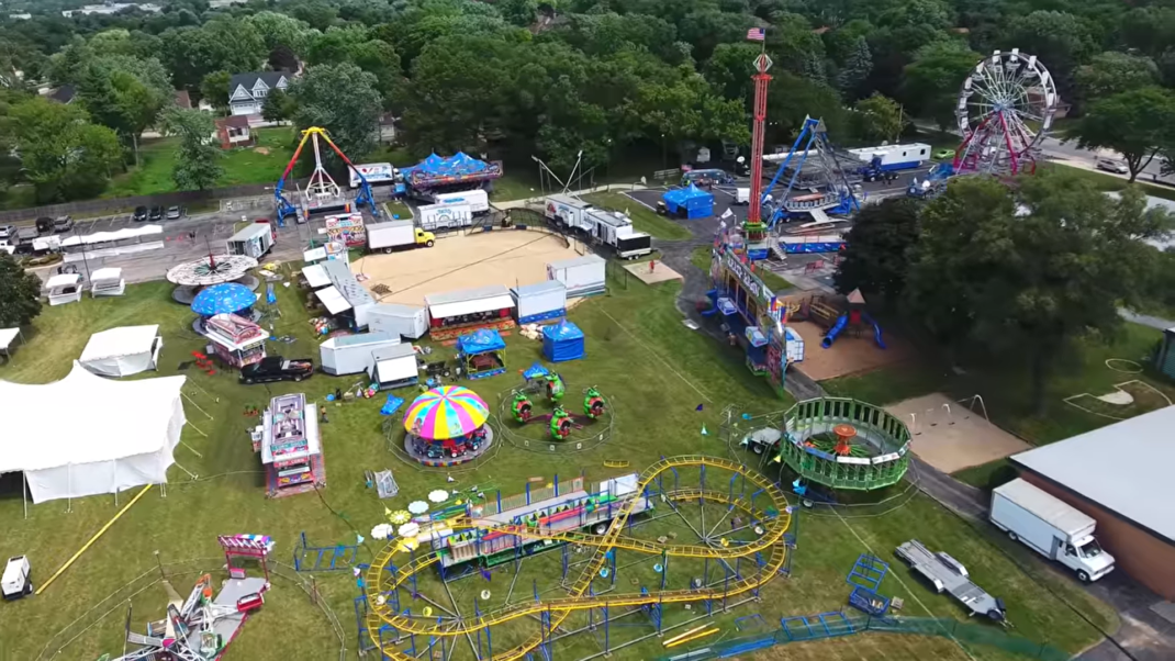 An aerial view of the festive grounds at Itasca Fest, colorful carnival rides, tents, and booths, set against a backdrop of green trees and residential houses.