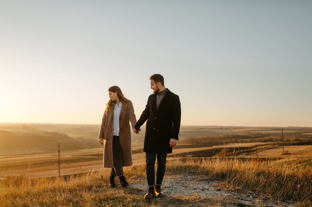 A couple holding hands and walking on a path overlooking vast rolling hills illuminated by a golden sunset.